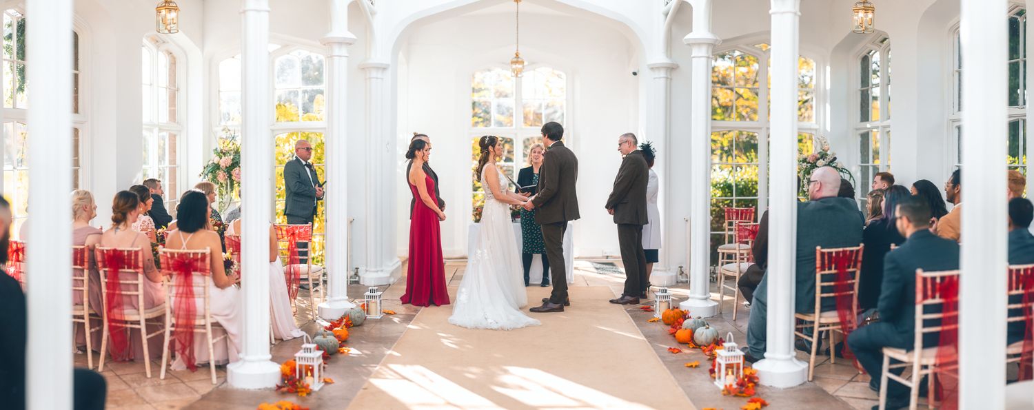 Wedding ceremony in a sunlit white glass orangery with autumn leaf decorations, lanterns and pumpkins along the aisle, bright venue ceremony photography St Audrie\