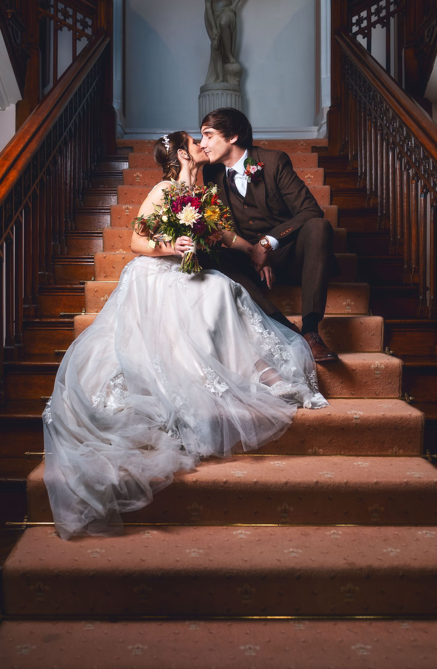 Bride and groom sharing a kiss seated on a grand carpeted staircase with colourful bouquet, romantic wedding photography St Audrie\