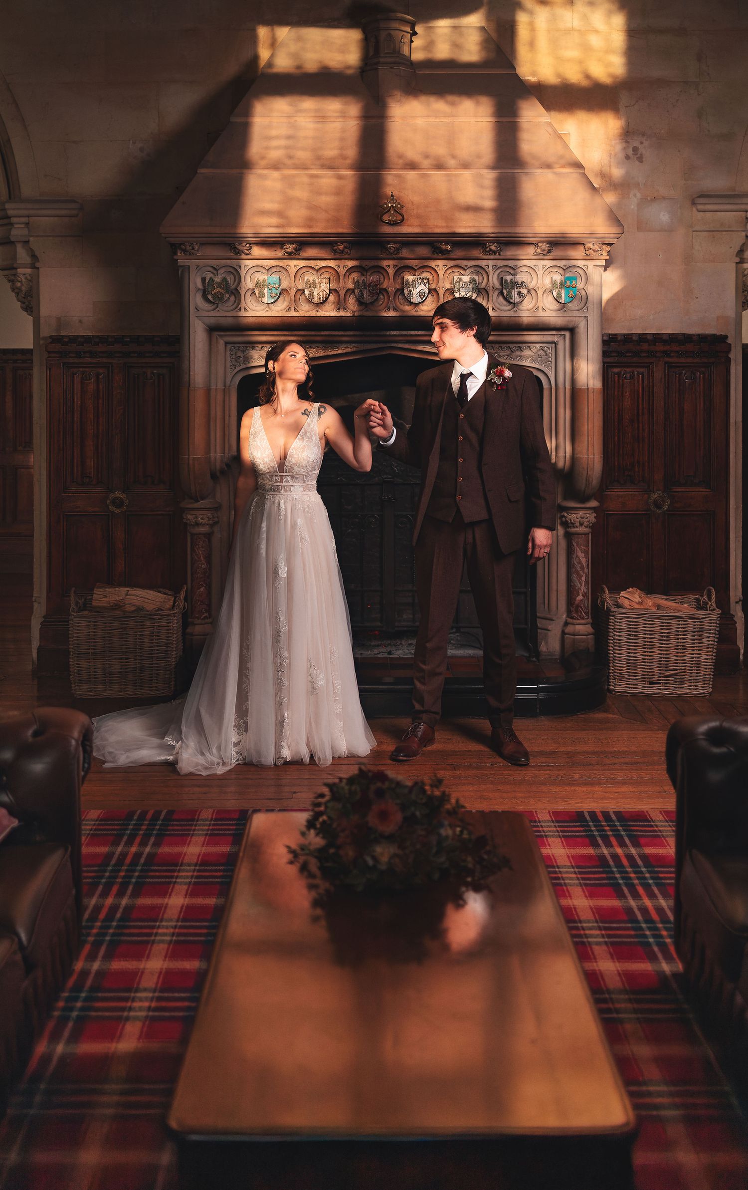 Bride and groom posing by an ornate stone fireplace in a grand hall with tartan table runner, elegant wedding portrait photography St Audrie\
