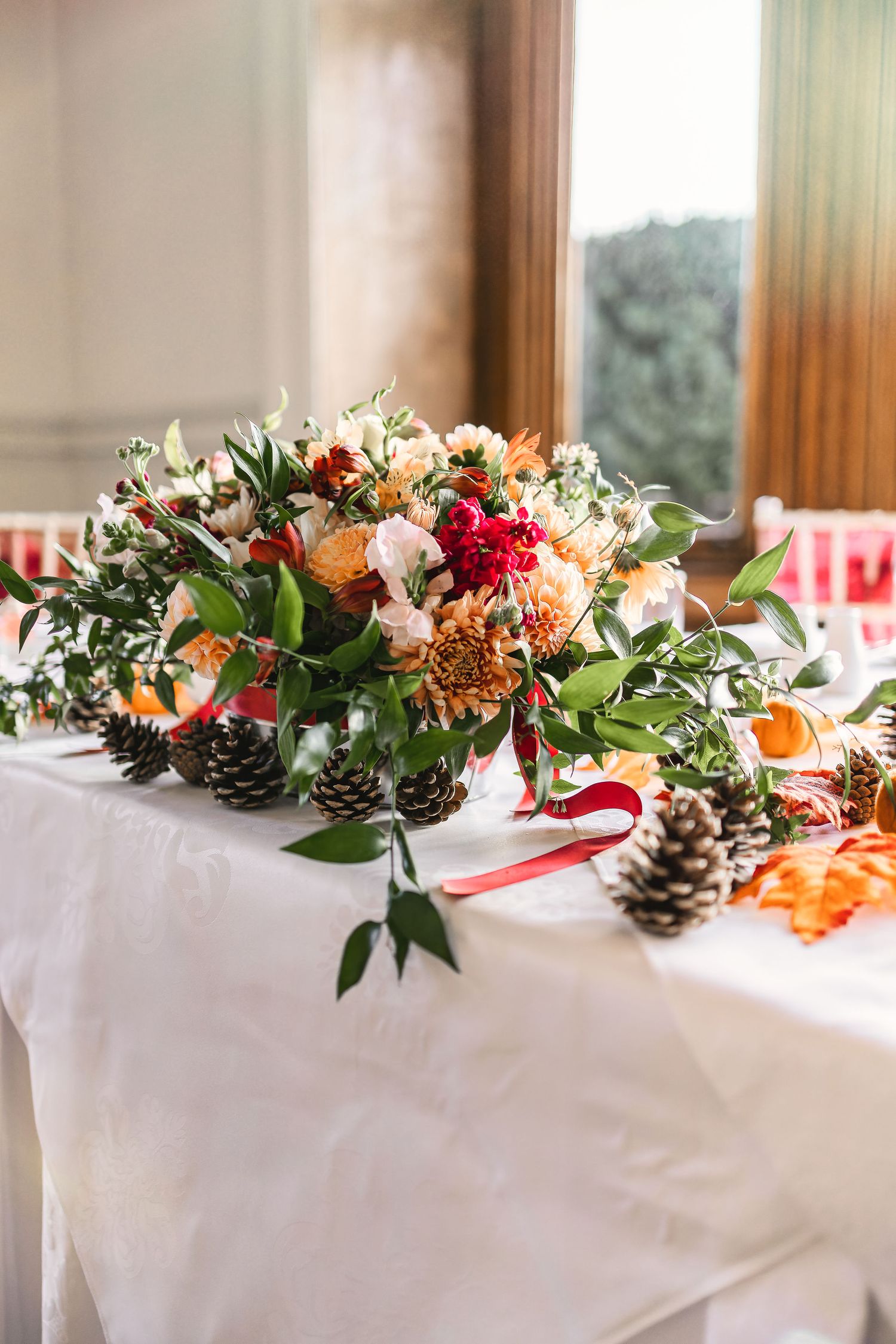 Autumn wedding table centrepiece with orange dahlias, red flowers, pine cones and greenery on a white tablecloth, wedding detail photography St Audrie\