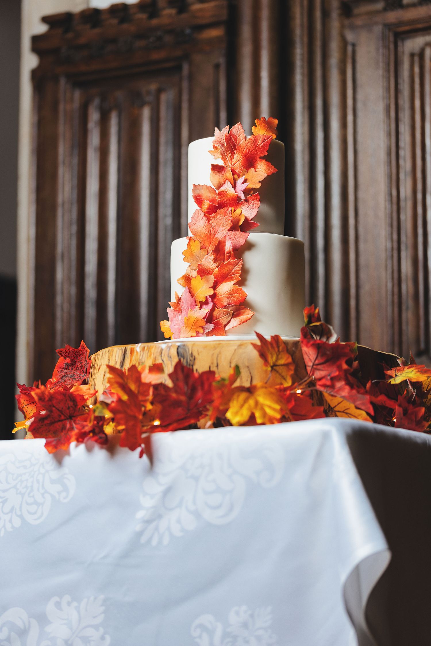 Three-tier white wedding cake decorated with cascading autumn leaves on a wooden log stand, wedding detail photography St Audrie\