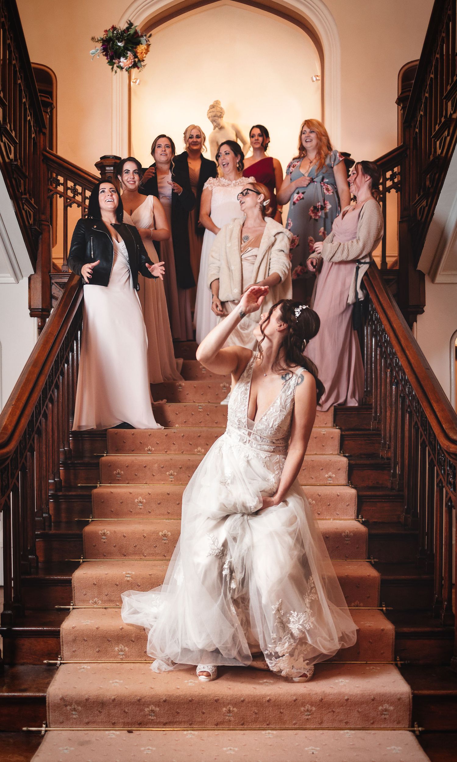 Bride on a grand wooden staircase with wedding party gathered above, elegant venue wedding photography St Audrie\