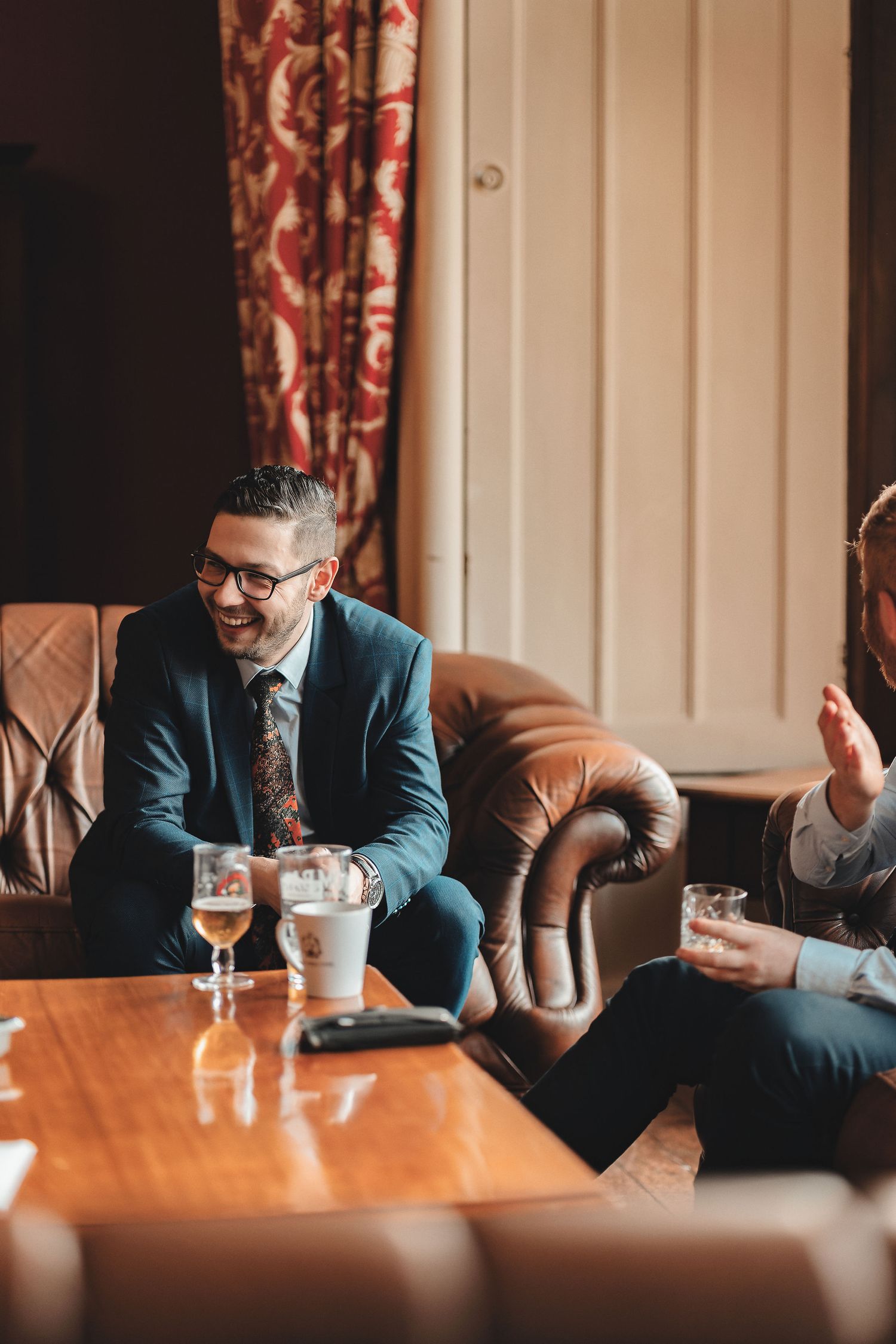 Groom in navy suit laughing with groomsman in leather Chesterfield chairs with drinks in a grand venue lounge, candid groom prep wedding photography St Audrie\