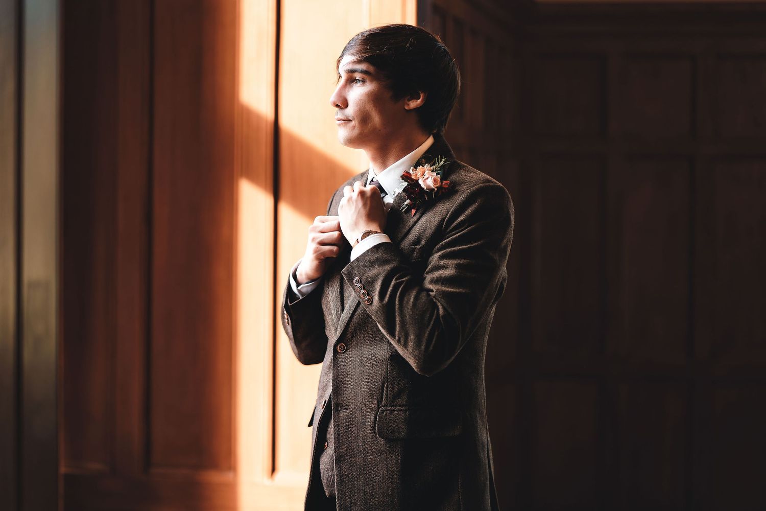 Groom in brown tweed suit adjusting his tie by a window with warm golden light and boutonniere, dramatic groom portrait wedding photography St Audrie\