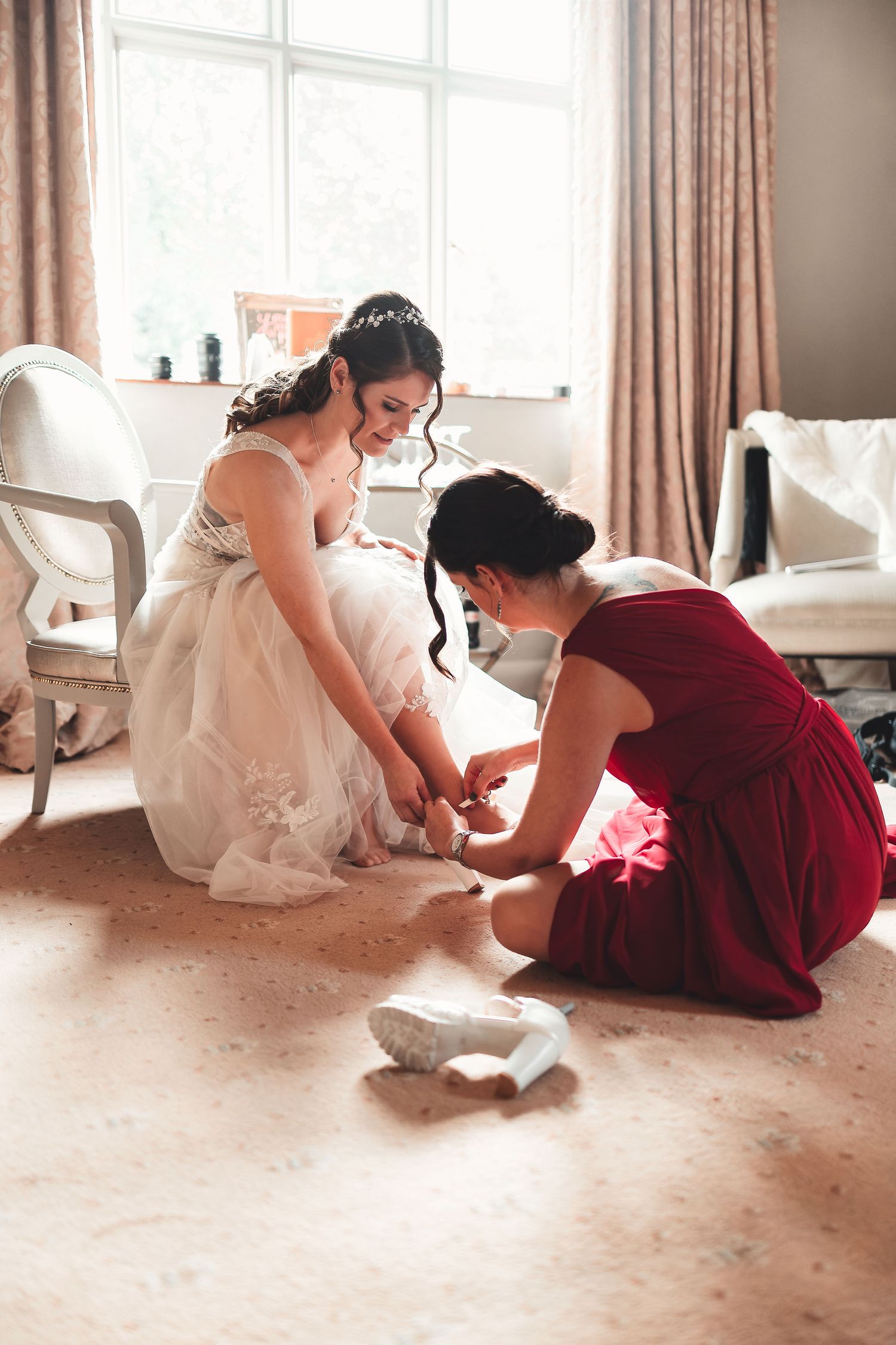 Bridesmaid in burgundy dress helping the bride put on shoes in a bright bridal suite with window light, candid bridal prep photography St Audrie\