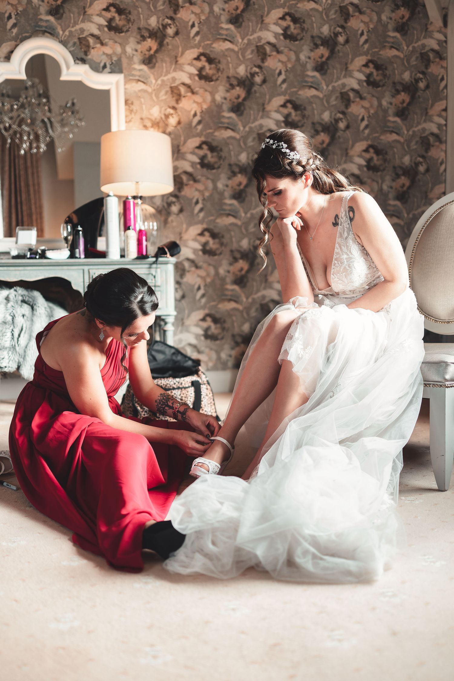 Bridesmaid in red dress helping the bride put on shoes in a bridal suite with floral wallpaper, candid bridal prep photography St Audrie\