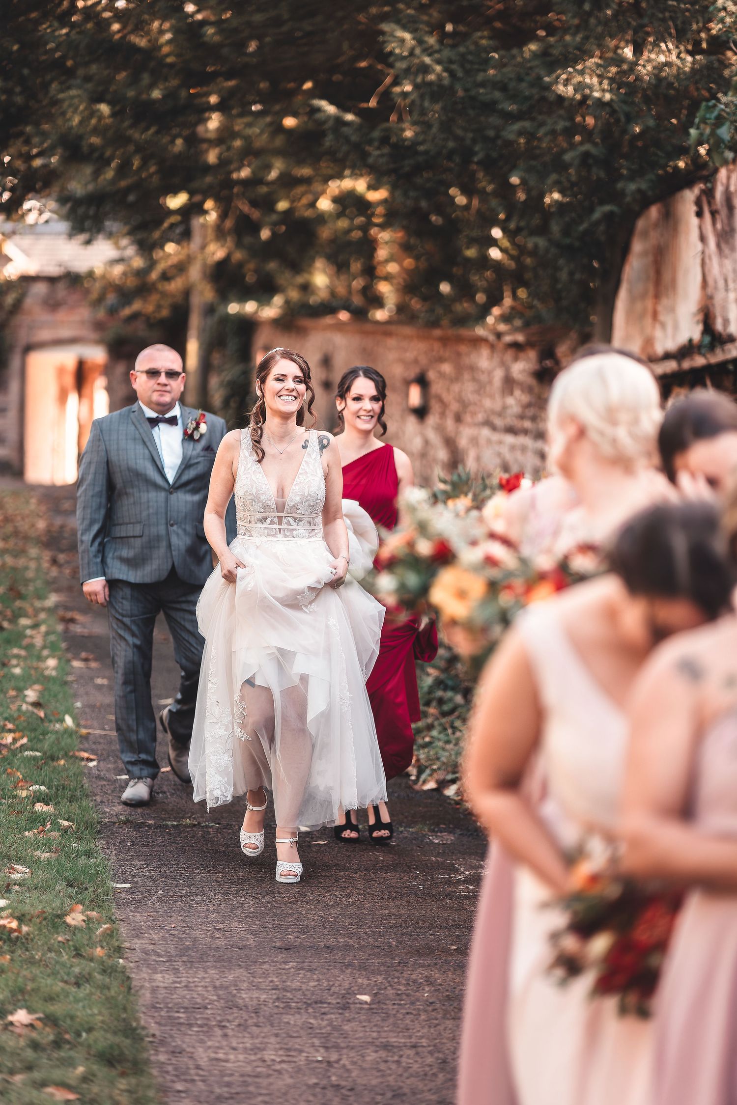 Bride walking along a path with her father and bridesmaids in pink and red dresses, candid wedding procession photography St Audrie\