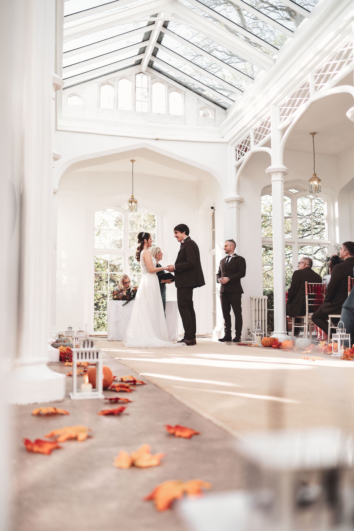 Wedding ceremony in a bright white glass orangery with autumn leaf decorations and lanterns along the aisle, autumn wedding ceremony photography St Audrie\