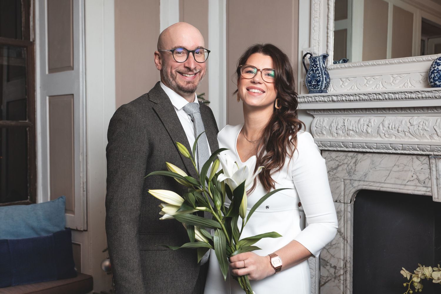 Bride and groom smiling together by a marble fireplace holding a lily bouquet indoors, intimate wedding portrait photography Southampton